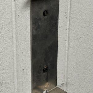 May include: A silver metal bracket fixed to a white wall. The bracket is rectangular with a top and two lower extensions. Screws attach the bracket to the wall. It is designed to hold an object, likely for storage or display.