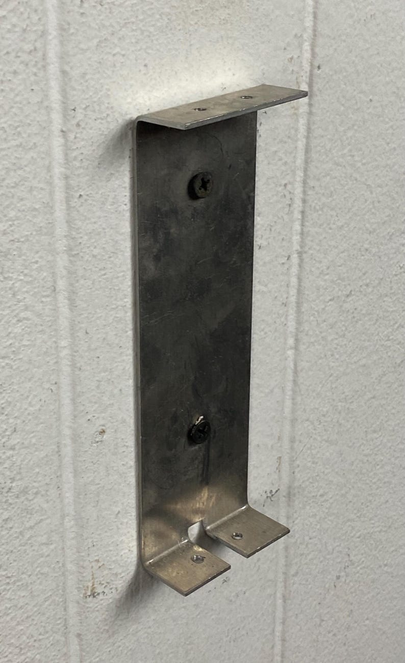 May include: A silver metal bracket fixed to a white wall. The bracket is rectangular with a top and two lower extensions. Screws attach the bracket to the wall. It is designed to hold an object, likely for storage or display.