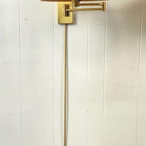 May include: A brass wall lamp with a cream lampshade. The lamp has a rectangular base, an adjustable arm, and a long, slender stem. The shade is a soft, warm colour, and the fixture is mounted on a white wall.