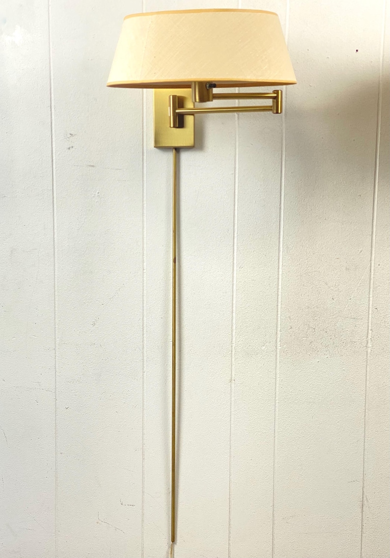May include: A brass wall lamp with a cream lampshade. The lamp has a rectangular base, an adjustable arm, and a long, slender stem. The shade is a soft, warm colour, and the fixture is mounted on a white wall.