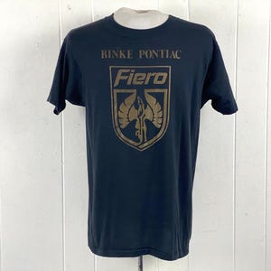 Puede incluir: Camiseta negra con un gráfico dorado que presenta el texto "RINKE PONTIAC" encima de "Fiero" y un diseño de escudo. La camiseta es de cuello redondo.