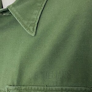 May include: Close-up of a green shirt, showcasing the collar and a pocket. The fabric has a textured appearance, and the stitching is visible along the collar and pocket edges. The shirt appears to be made of a durable material.