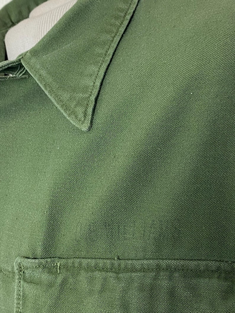 May include: Close-up of a green shirt, showcasing the collar and a pocket. The fabric has a textured appearance, and the stitching is visible along the collar and pocket edges. The shirt appears to be made of a durable material.