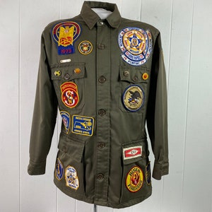 Op de afbeelding: Een groene utility-jas met meerdere patches, waaronder een "Ruger Firearms"-patch, een "CCI"-patch en een "Police"-patch. De jas heeft een knoopsluiting en twee grote zakken.