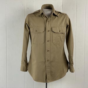 May include: A tan-colored button-down shirt with two chest pockets and long sleeves. The shirt has a classic collar and epaulets on the shoulders. The shirt is displayed on a mannequin against a white background.