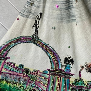 May include: A white fabric skirt with a colorful, sequined design of a city skyline and an archway. The design features a woman holding a globe on top of the archway.