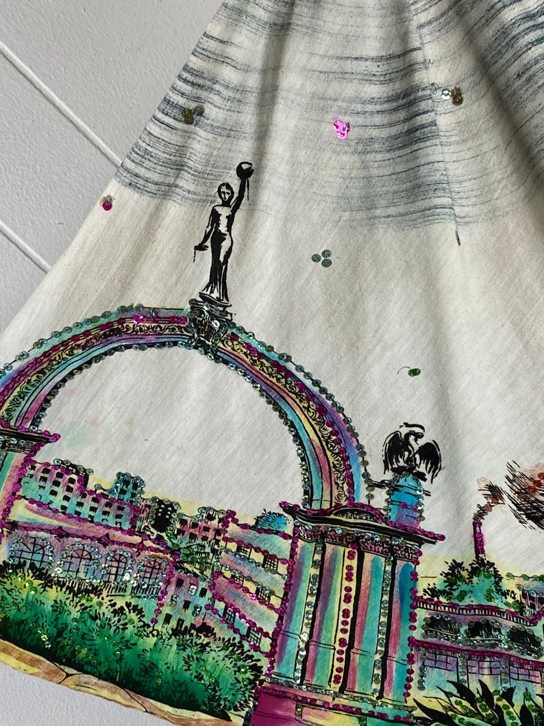 May include: A white fabric skirt with a colorful, sequined design of a city skyline and an archway. The design features a woman holding a globe on top of the archway.