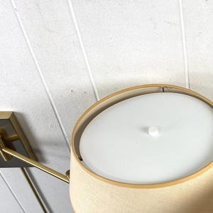May include: A contemporary wall lamp featuring a beige lampshade and a gold-toned metal arm and base. The lampshade has a white diffuser. The lamp is mounted on a white wall.