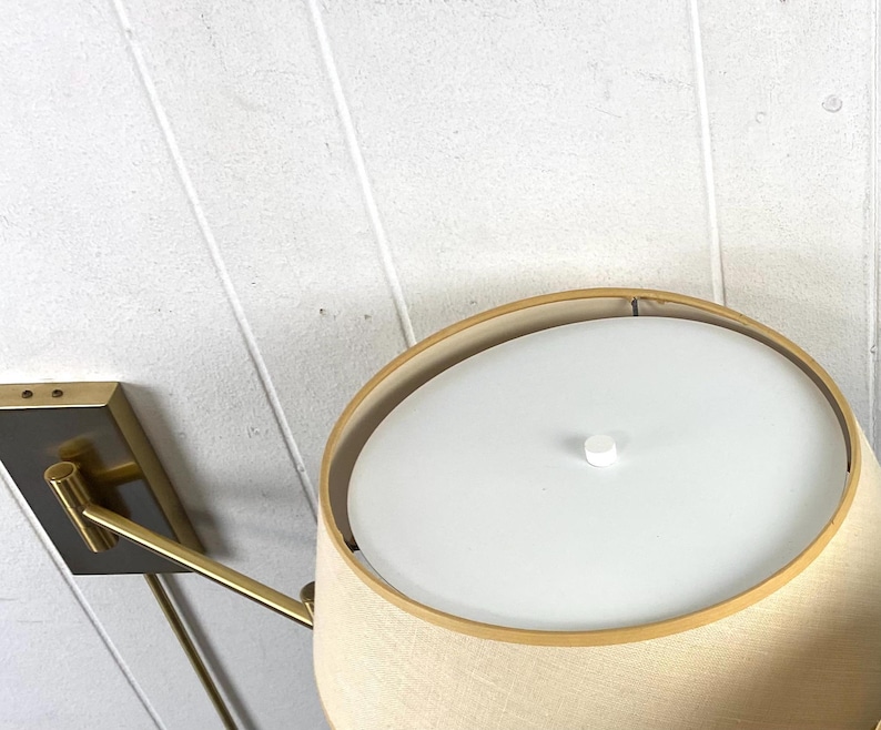 May include: A contemporary wall lamp featuring a beige lampshade and a gold-toned metal arm and base. The lampshade has a white diffuser. The lamp is mounted on a white wall.