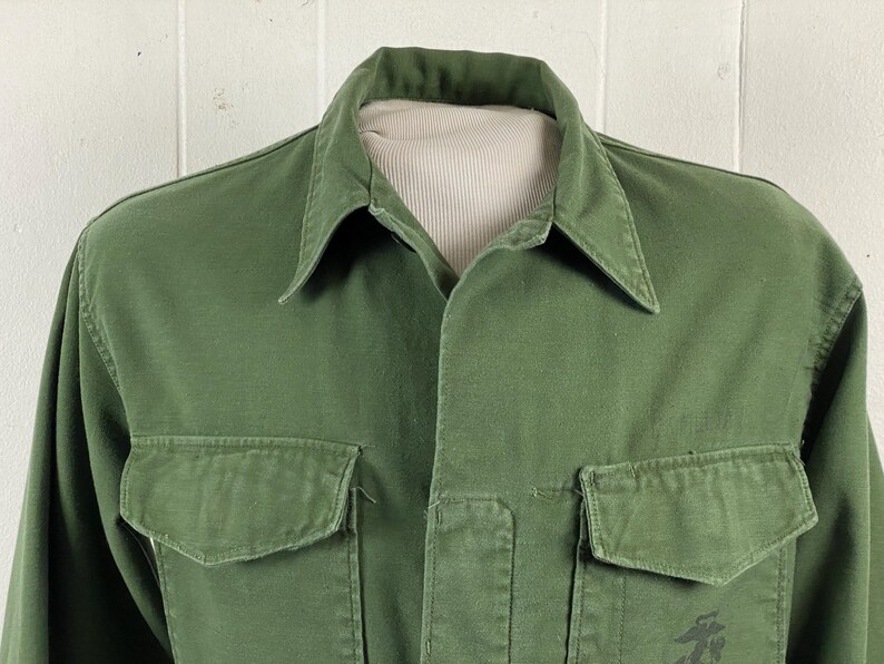 May include: Olive green button-up shirt with two front pockets and a collared neckline. The shirt is displayed on a mannequin. The shirt has a small graphic on the lower right side.