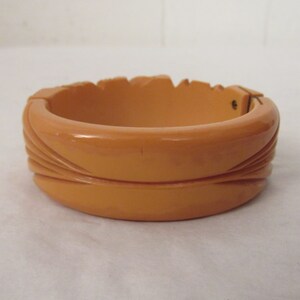 Puede incluir: Un brazalete vintage de baquelita de color naranja-marr&oacute;n con un dise&ntilde;o tallado. El brazalete tiene un cierre de presi&oacute;n.