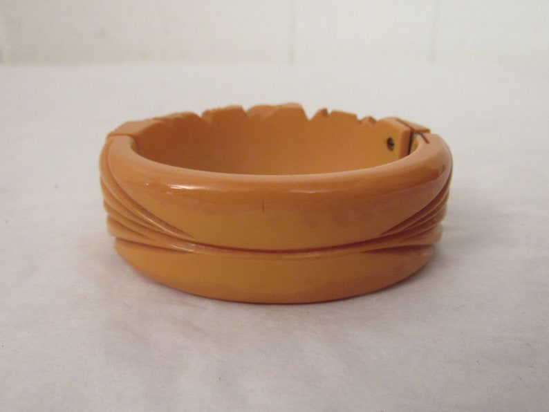 Puede incluir: Un brazalete vintage de baquelita de color naranja-marr&oacute;n con un dise&ntilde;o tallado. El brazalete tiene un cierre de presi&oacute;n.
