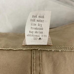May include: Close-up of a beige fabric with a sewn seam and a care label. The label reads "Hand wash, Cold water, Line dry, Preshrunk, May shrink additional 3-5%."