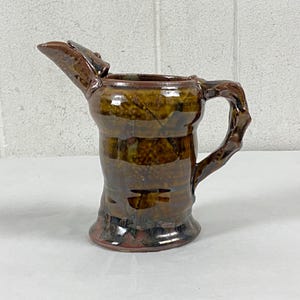 May include: A handcrafted ceramic pitcher with a unique, textured design. The pitcher features a dark brown glaze with lighter brown and green accents. It has a spout, a handle, and a bulbous body. The pottery is suitable for serving beverages.