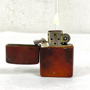Puede incluir: Un encendedor vintage de metal marrón, abierto y con llama. El encendedor tiene un cuerpo rectangular con bordes redondeados y una tapa con bisagras. La superficie muestra signos de desgaste, con un diseño descolorido en el frente.