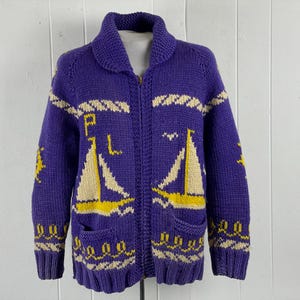May include: A purple knitted cardigan with a shawl collar and a zipper closure. The sweater features cream-colored sailboat and wave designs, with the letters "P" and "L" on the front. The sleeves have yellow star accents.