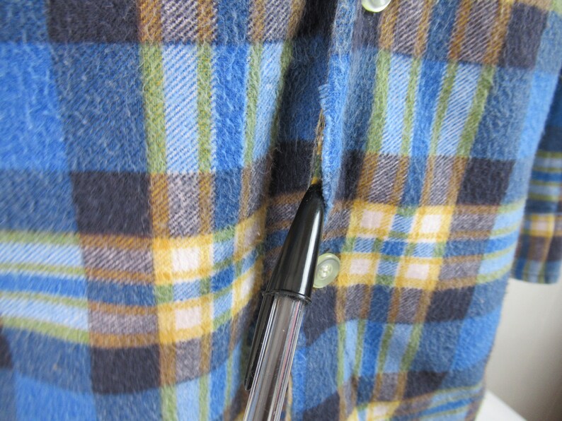 以下が含まれることがあります： 青、黄、茶色、白の格子柄のフランネルシャツのクローズアップ。シャツはボタン留めフロントと長袖です。