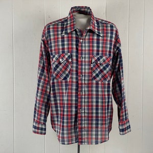 Puede incluir: Una camisa de manga larga con botones y un estampado de cuadros rojo, blanco y azul. La camisa tiene dos bolsillos en el pecho con cierre de botón.