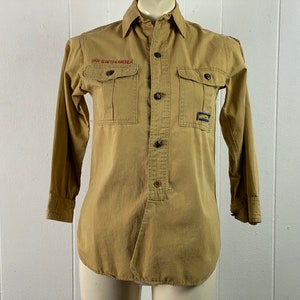 May include: A tan long-sleeved button-down shirt with two large pockets on the front. The shirt has "BOY SCOUTS OF AMERICA" embroidered on the left pocket and a patch on the right pocket.