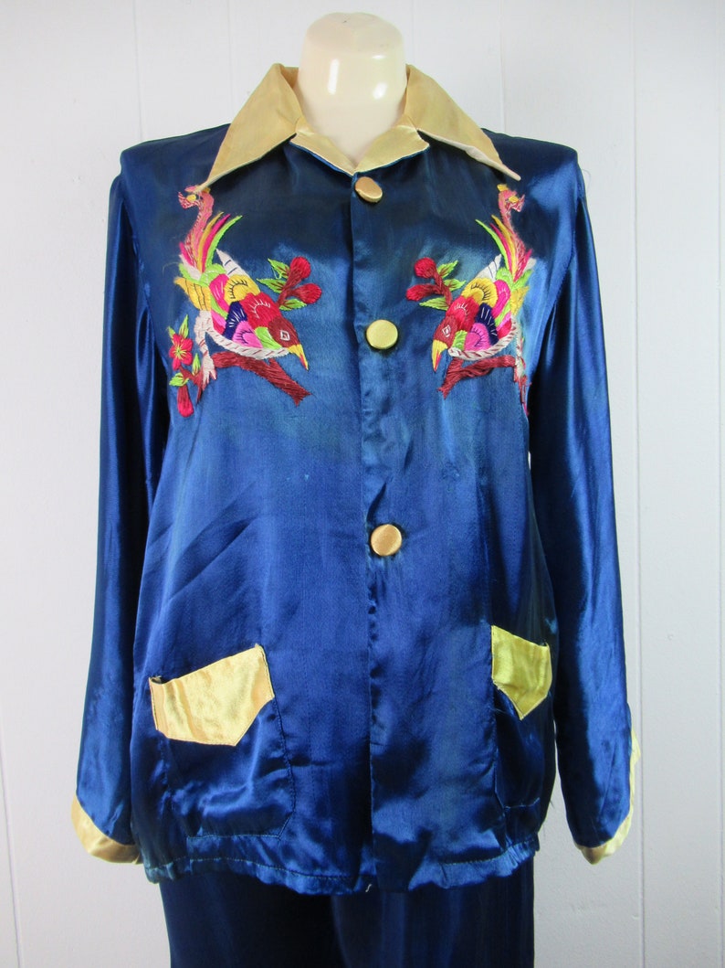 May include: A blue satin pajama set with gold trim and embroidered birds. The jacket has a collar, button closure, and two pockets. The pants have a straight leg.