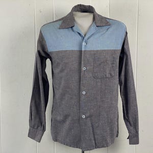 Puede incluir: Camisa abotonada de manga larga con un diseño de dos tonos. La sección superior de la camisa y el cuello son de color azul claro, mientras que el resto de la camisa es de color gris oscuro. Tiene un bolsillo en el pecho y puños abotonados.