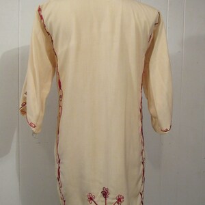 May include: A cream-colored tunic with red embroidery. The embroidery is in a floral pattern and is on the neckline, sleeves, and hem of the tunic.
