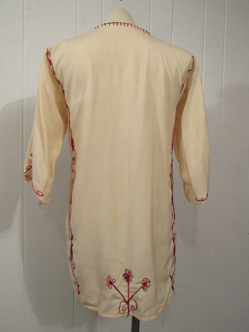 May include: A cream-colored tunic with red embroidery. The embroidery is in a floral pattern and is on the neckline, sleeves, and hem of the tunic.
