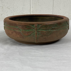 May include: A terracotta planter with a dragonfly design in green glaze. The planter is round and has a wide rim.
