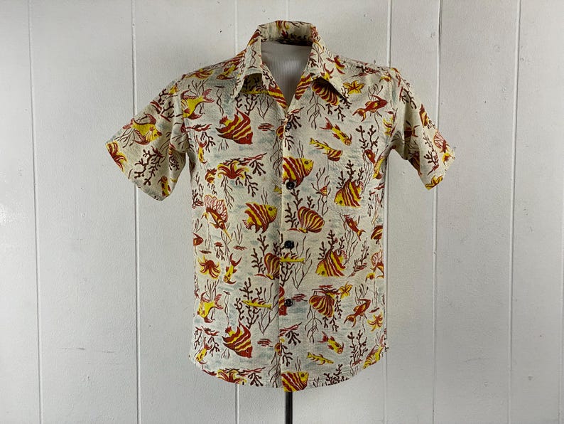 May include: A short-sleeved button-down shirt with a white background and a colorful print of fish and coral. The shirt has a collar and a button-up front.