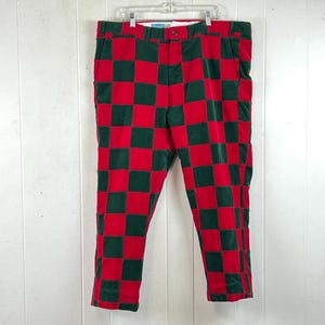 May include: A pair of trousers featuring a bold red and dark green checkered pattern. The pants have a structured waistband with a button closure, a front zipper fly, and belt loops. The fabric appears to have a textured, corduroy-like finish.