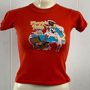 May include: A red t-shirt featuring a pirate-themed graphic. The graphic includes a pirate with a pistol, a treasure chest, and the words "Pirate's Gold Jamaica". The shirt is displayed on a mannequin.