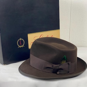 Op de afbeelding: Een bruine fedora hoed met een donkerbruin lint en een veermotief. De hoed staat naast een zwarte doos met gouden letters en een logo. De hoed heeft een klassiek ontwerp.