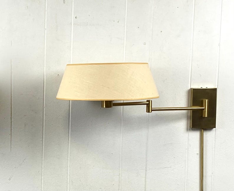 May include: A wall-mounted lamp featuring a beige lampshade and a brass-coloured adjustable arm. The lamp is mounted on a rectangular brass-coloured base. The lamp is plugged into an electrical outlet.