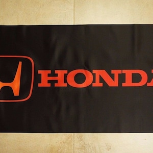 Puede incluir: Bandera negra con el logotipo de Honda en naranja y rojo. El logotipo es una 'H' estilizada dentro de un cuadrado, con la palabra 'HONDA' en letras rojas y en negrita. Hay ojales en cada esquina.