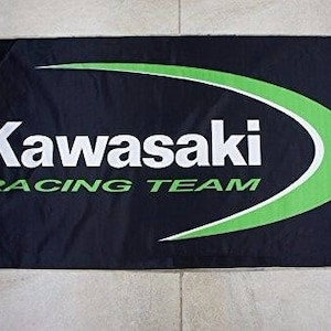 Puede incluir: Bandera negra y verde con un logotipo blanco de Kawasaki Racing Team. El logotipo está en una fuente blanca sans-serif con un swoosh verde detrás.