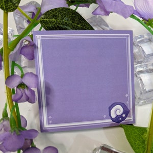 May include: A purple note card with a white border and a small star design. The card has a white border and a small star design. The card has a small, white, crescent moon design in the bottom right corner.