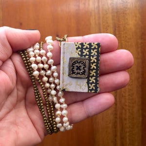 May include: A gold chain necklace with a small book charm. The book is covered in black and gold patterned fabric with a gold square accent. The chain is layered with a pearl and a gold bead necklace.