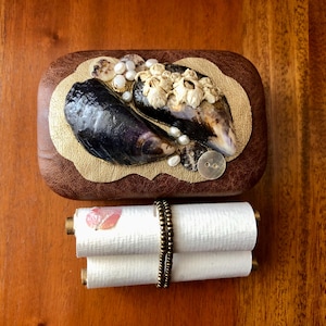May include: A small wooden box with a gold-coloured lid decorated with seashells, pearls, and a button. The box is closed and sitting on a wooden surface. A white scroll with a pink flower design is tied with a brown cord and is sitting below the box.