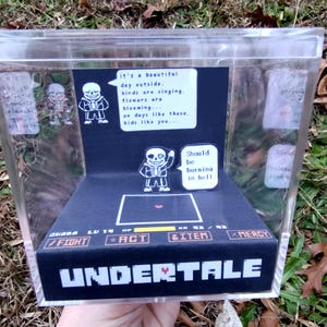 May include: A clear acrylic display case featuring a diorama inspired by the video game Undertale. The scene includes a character illustration, text bubbles, and the game's interface. The text reads "Should be burning in hell."