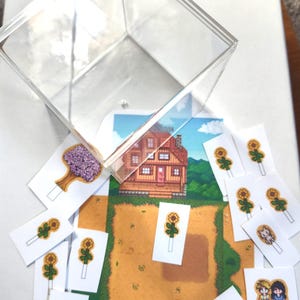 May include: A clear acrylic box sits above a collection of game-themed cards. The cards feature pixelated graphics of a house, farm, sunflowers, and characters. The farm name and date are visible on one card.