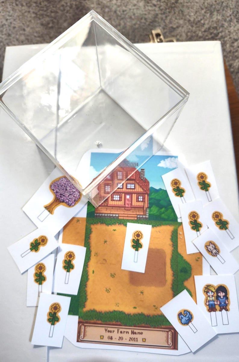 May include: A clear acrylic box sits above a collection of game-themed cards. The cards feature pixelated graphics of a house, farm, sunflowers, and characters. The farm name and date are visible on one card.