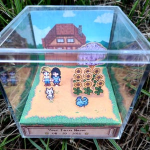 May include: A pixel art diorama inside a clear acrylic box. The scene depicts a farm with a house, sunflowers, and characters. The base of the box has text that reads "Your Farm Name" and a date. The diorama is set on a grassy surface.