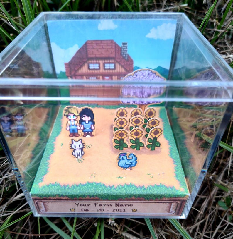 May include: A pixel art diorama inside a clear acrylic box. The scene depicts a farm with a house, sunflowers, and characters. The base of the box has text that reads "Your Farm Name" and a date. The diorama is set on a grassy surface.