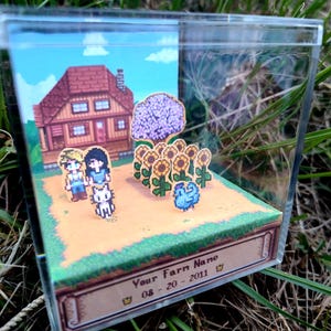 May include: A miniature diorama featuring pixelated characters, a house, sunflowers, and a blue creature. The scene is set in a clear acrylic box, with the text "Your Farm Name" and a date on the base.