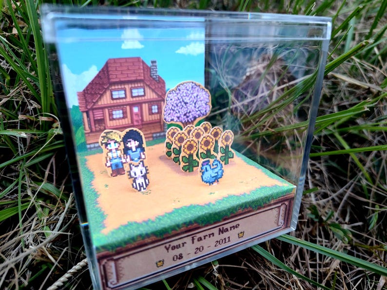 May include: A miniature diorama featuring pixelated characters, a house, sunflowers, and a blue creature. The scene is set in a clear acrylic box, with the text "Your Farm Name" and a date on the base.