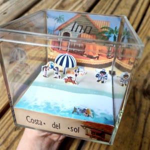 May include: A clear acrylic box diorama featuring a beach scene with miniature figures, a striped umbrella, and a building labeled "Inn." The base of the diorama reads "Costa del sol."