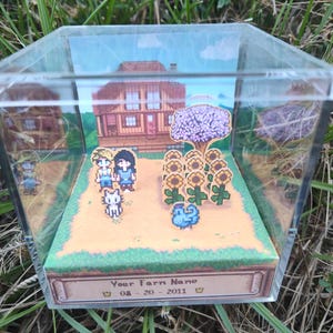 May include: A clear acrylic display case featuring a pixelated diorama of a farm scene. The scene includes a house, sunflowers, characters, and the text "Your Farm Name" with a date. The diorama is set against a backdrop of a blue sky and green grass.