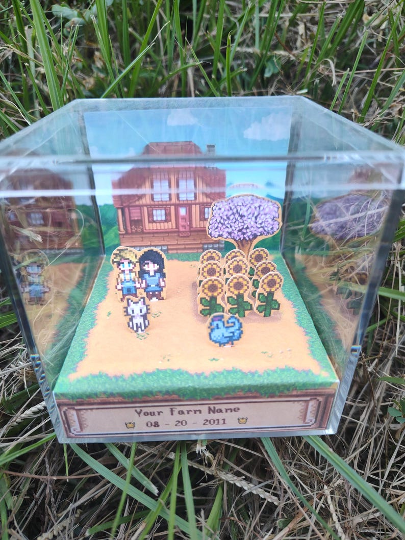 May include: A clear acrylic display case featuring a pixelated diorama of a farm scene. The scene includes a house, sunflowers, characters, and the text "Your Farm Name" with a date. The diorama is set against a backdrop of a blue sky and green grass.