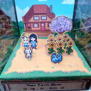 May include: A diorama featuring a pixelated farm scene with a house, sunflowers, and characters. The scene includes a cat, a blue bird, and two people. The base of the diorama has text that reads "Your Farm Name" and a date.