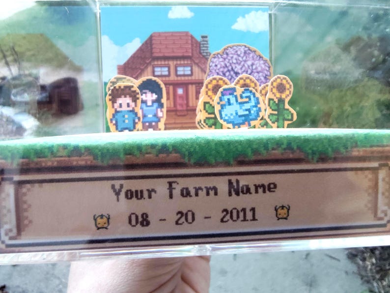 May include: A pixelated diorama featuring a farm scene with a house, characters, and sunflowers. The text "Your Farm Name" and the date "08-20-2011" are displayed on a banner at the bottom. The scene is set inside a clear display case.